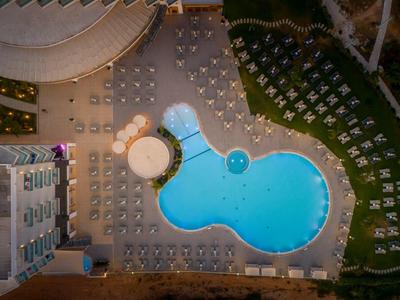 Blick von oben: Großer Pool in ungewöhnlicher Form, umgeben von Sonnenliegen und Gebäuden.