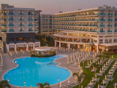 Großes Hotel mit mehreren Stockwerken, runden Pool und Reihen von Liegestühlen bei Sonnenuntergang.