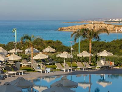 Pool mit Liegestühlen und Sonnenschirmen vor Palmen, grüner Küste und blauem Meer im Hintergrund.
