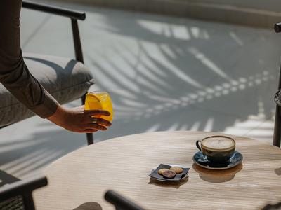 Personne tenant un verre avec une boisson orange près d'une table avec une tasse et une collation dans une pièce lumineuse.
