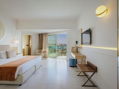 Chambre d'hôtel lumineuse avec grand lit, TV, bureau et balcon avec vue sur la mer.