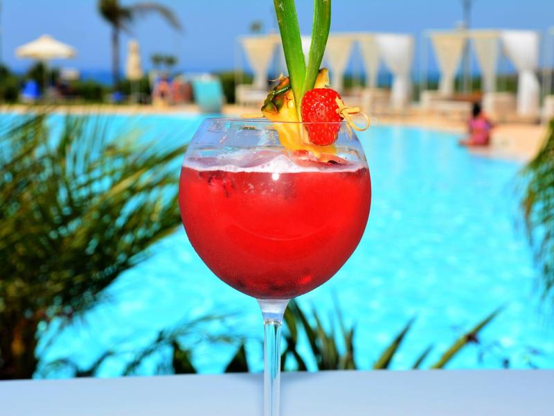Rotes Cocktailglas mit Fruchtgarnitur vor blauem Pool und sommerlicher Umgebung.