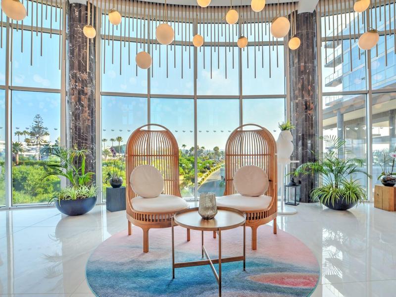 Heller Raum mit großen Glasfenstern, zwei Korbsesseln, blau-rosa Teppich und Pflanzen.