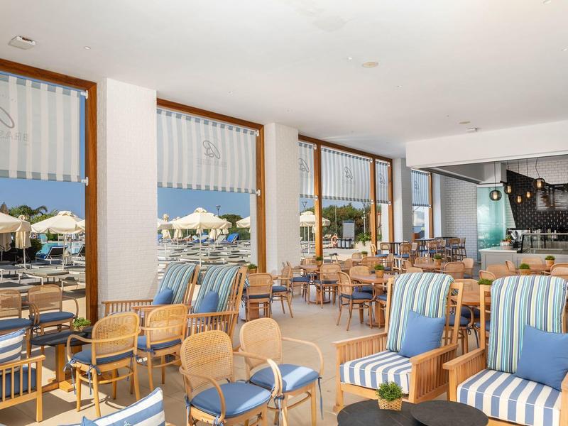 Helles Café mit blauen und weißen Sesseln, großen Glasfenstern und Blick auf Terrasse mit Sonnenschirmen.