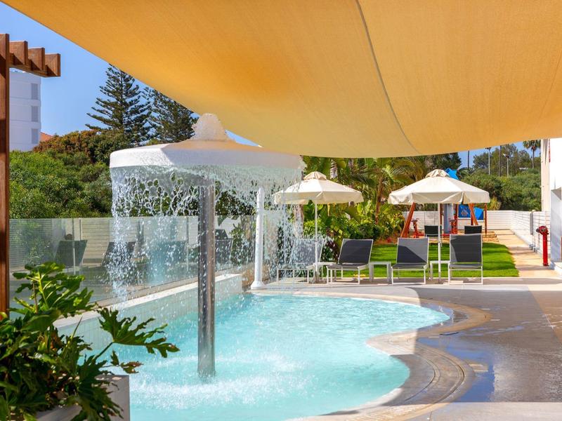 Schwimmbad mit Wasserfall, Sonnenschirmen, Liegestühlen und Sonnensegel unter Bäumen.