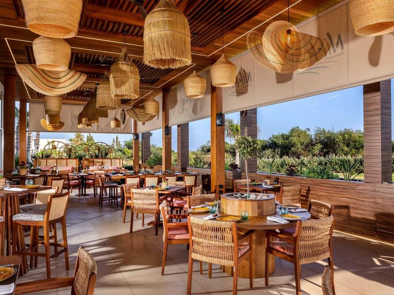 Großes, offenes Restaurant mit Holzmöbeln, vielen Lampen aus Rattan und Blick auf grüne Pflanzen.