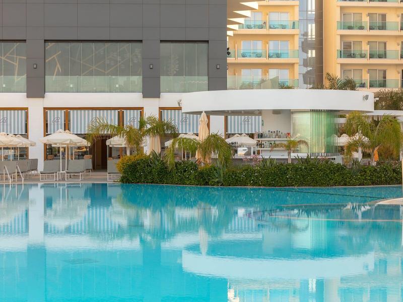 Großer blauer Pool vor moderner Hotelterrasse mit weißen Sonnenschirmen und grünen Pflanzen.
