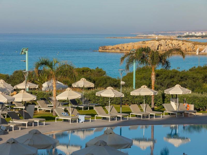 Pool mit Liegestühlen und Sonnenschirmen vor Palmen, grüner Küste und blauem Meer im Hintergrund.