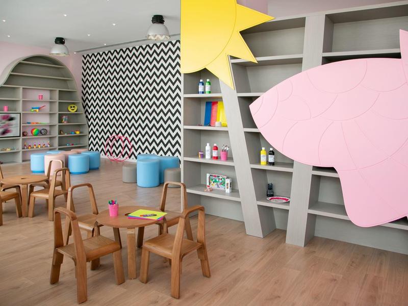 Kindergartenraum mit Holztischen, Stühlen, bunten Regalen und einem großen gelben Stern an der Wand.