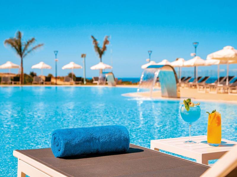 Poollandschaft mit blauem Wasser, Liegestuhl, blauem Handtuch und orangem Cocktail bei Sonnenschein.