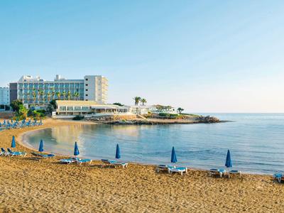 Pláž s modrými slunečníky, lehátky a hotelovou budovou u moře za jasné oblohy.