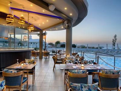 Offenes Hotelrestaurant mit Tischen und Stühlen, Blick auf Wasser und Hafen bei Sonnenuntergang.