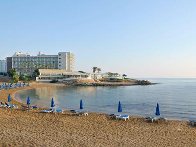Ein ruhiger Sandstrand mit blauen Sonnenschirmen vor einem Hotel am Meer bei klarem Himmel.