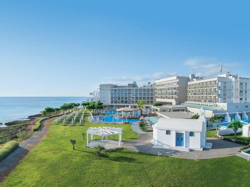 Luxe hotel aan zee met zwembad en groen gazon onder blauwe lucht.