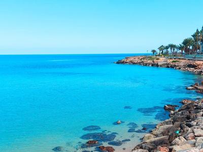 Costa con acqua azzurra limpida, rocce e edifici lungo la riva sotto un cielo azzurro.