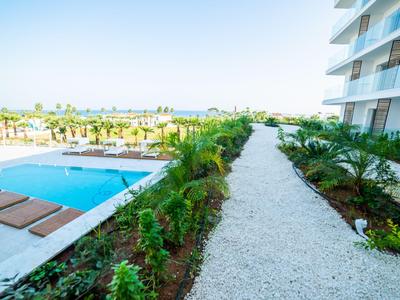 Un sentiero curato con piante corre accanto a un hotel, con una piscina e lettini a sinistra sotto un cielo azzurro.