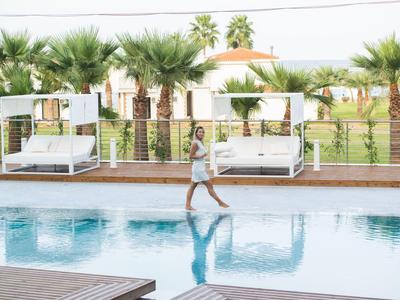 Donna che cammina a piedi nudi lungo il bordo della piscina con palme e lettini sullo sfondo.