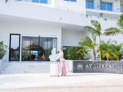 Donna con valigia davanti a un edificio di hotel moderno con piante e sole.