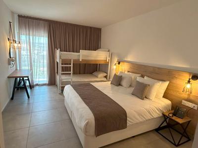 Hell eingerichtetes Hotelzimmer mit Doppelbett, Etagenbett und Balkon mit Vorhängen.