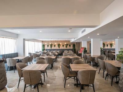 Heller, moderner Speisesaal mit vielen Holzstühlen und Tischen, großen Fenstern und Pflanzen.