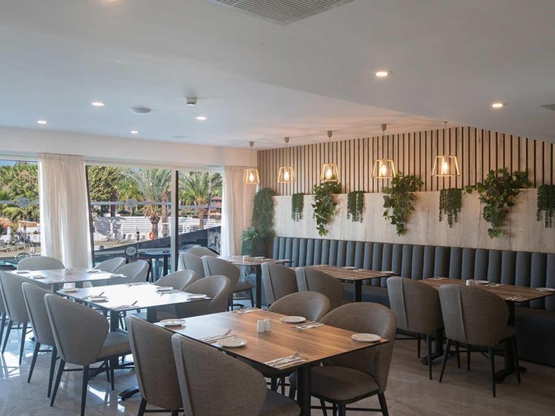 Helles Restaurant mit grauen Stühlen, Holztischen und grünen Pflanzen an der Wand unter Deckenlichtern.