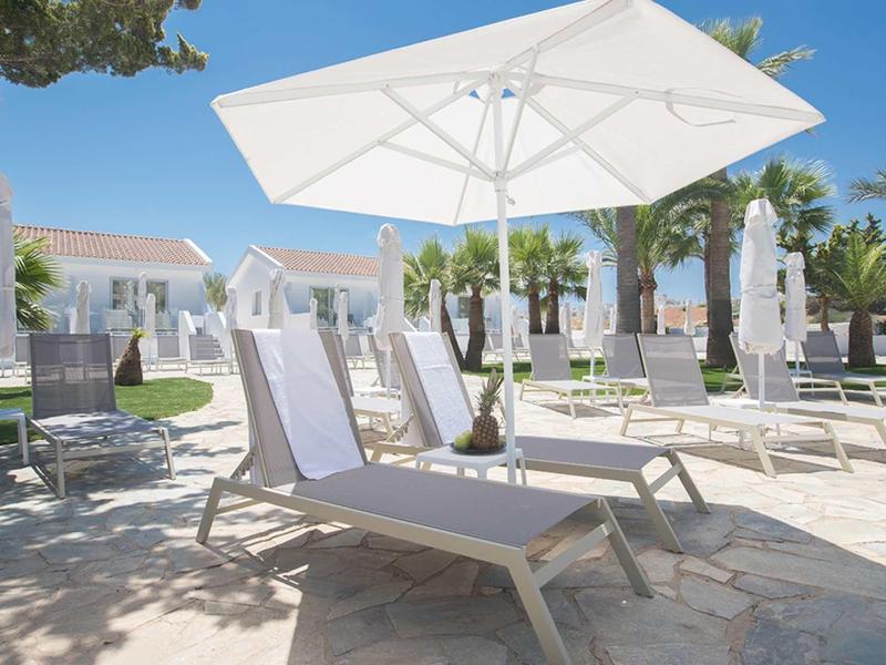 Mehrere weiße Liegestühle unter großen Sonnenschirmen auf gepflasterter Terrasse mit Bäumen.