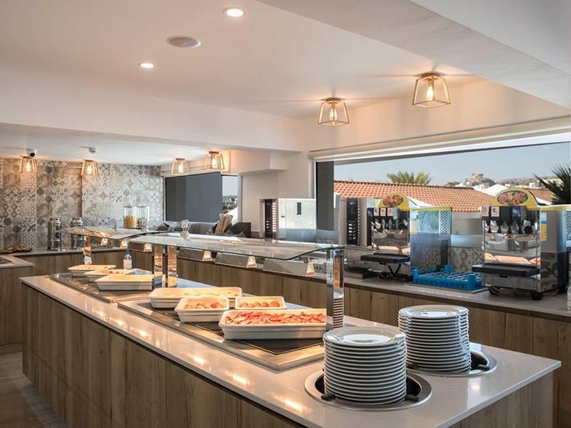 Moderne, helle Cafeteria mit Buffet, warmen Holztönen und weißer Decke, großes Fenster mit Außenblick.