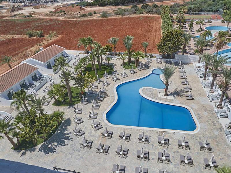 Großer Außenpool in tropischer Anlage mit Sonnenliegen und Palmen, umgeben von Feldern und Gebäuden.