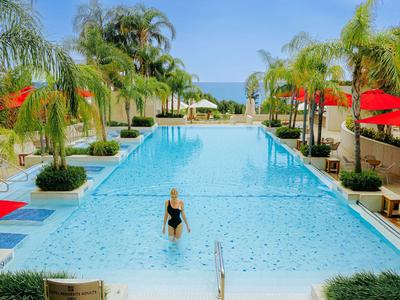 Mujer con traje de baño negro en una gran piscina al aire libre, rodeada de palmeras y sombrillas rojas.