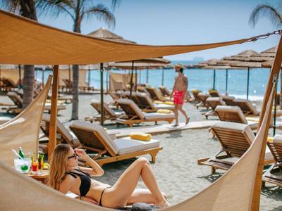 Mujer se relaja en hamaca en la playa con tumbonas y palmeras al fondo.