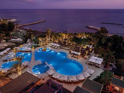 Großer, beleuchteter Pool mit Liegestühlen und Palmen an einem Hotel am Meer bei Dämmerung.
