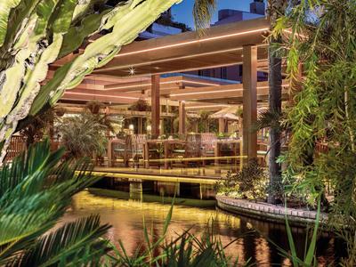 Moderner Hotelaußenbereich mit Wasser, Holzdeck und üppiger Bepflanzung bei Dämmerung.