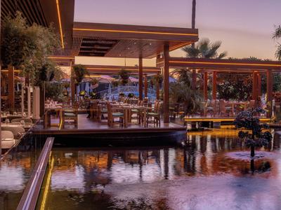 Restaurantterrasse mit Holztischen und Stühlen neben einem Wasserbecken bei Dämmerung.
