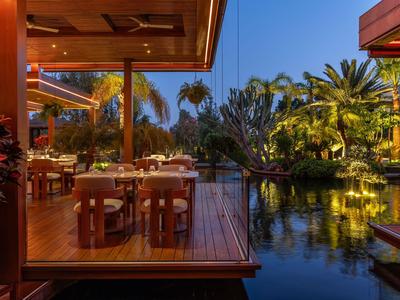 Abendliche Außenrestaurantterrasse mit Tischen neben Wasser und tropischer Vegetation.