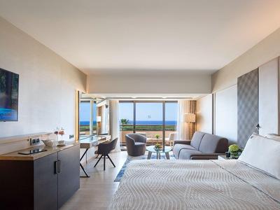 Modern hotel room with two beds, sitting area, and ocean view through large windows.