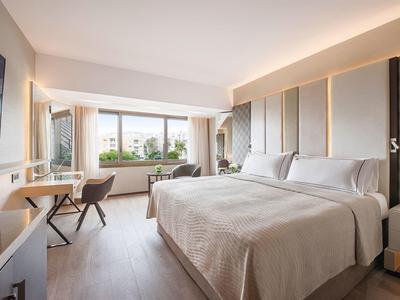 Bright hotel room with large bed, desk, and city view through a large window.
