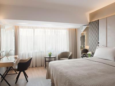 Modernes Hotelzimmer mit großem Bett, Schreibtisch und zwei Sesseln vor einem großen Fenster.