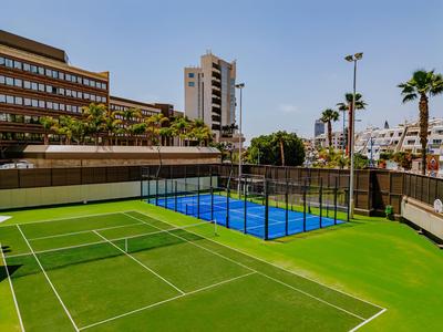 Hotel mit Tennis- und Paddle-Tennisplatz vor modernen Gebäuden unter blauem Himmel.