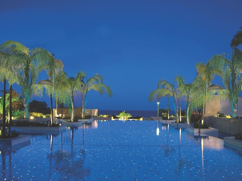 Illuminated hotel pool at night with palm trees and clear night sky.