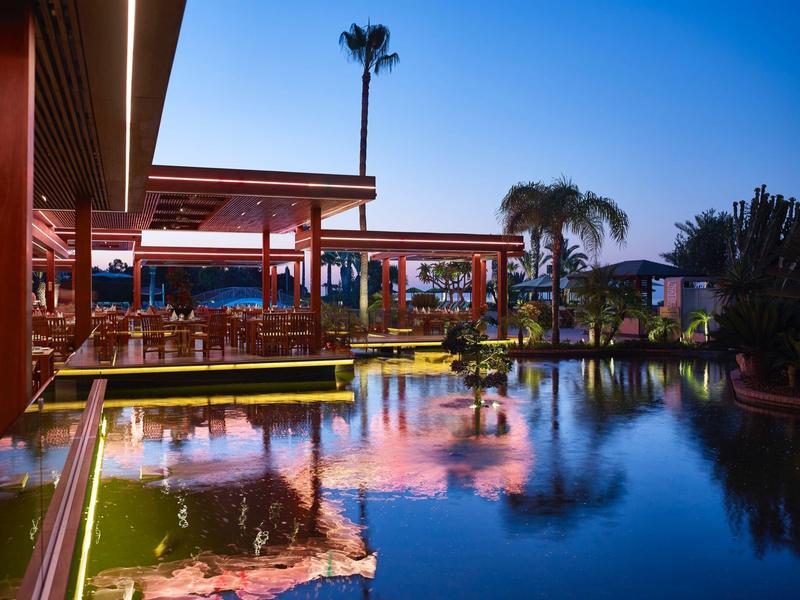 Outdoor restaurant by water with tables, chairs, and palm trees under clear sky