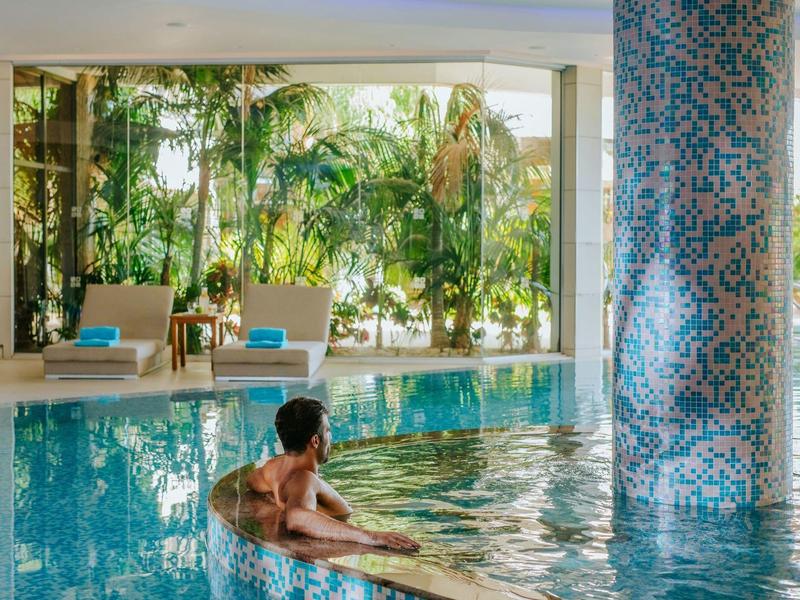 Man relaxes in indoor pool with blue tiled column and garden view.