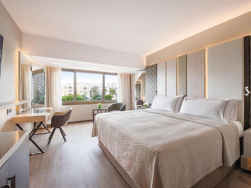 Bright hotel room with large bed, desk, and city view through a large window.