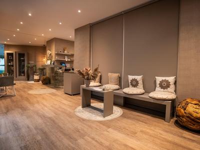 Gemütlicher Raum mit Holzboden, grauer Sofa-Ecke, Kissen, Pouf und offener Küche im Hintergrund.