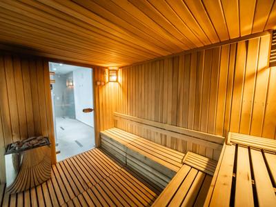 Innenraum einer Sauna mit Holzbänken, Holzwänden und warmem Licht. Türen öffnen in einen hellen Raum.