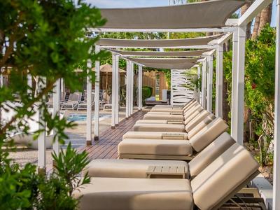 Longue rangée de chaises longues confortables sous une pergola blanche, entourée de plantes vertes.