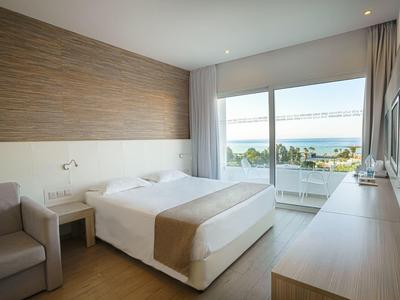 Helles, modernes Hotelzimmer mit Doppelbett, hellen Holzmöbeln und Meerblick durch Glasfenster.