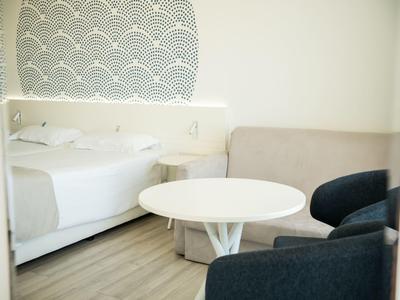 Kleines Zimmer mit rundem weißen Tisch, grauem Sessel, beige Couch und weißem Bett mit Kissen.