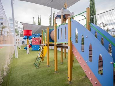 Bunter Kinderspielplatz mit Kletternetz, Rutschen und Trampolin unter Dach auf grünem Boden.