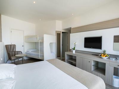 Helles Schlafzimmer mit Doppelbett, Etagenbett, TV, Sesseln und modernem Schrank in neutralen Tönen.