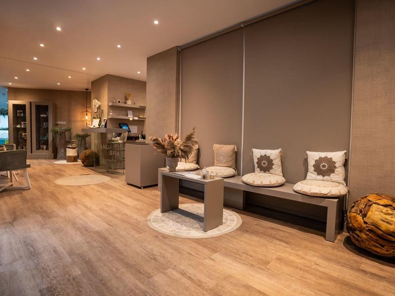 Gemütlicher Raum mit Holzboden, grauer Sofa-Ecke, Kissen, Pouf und offener Küche im Hintergrund.